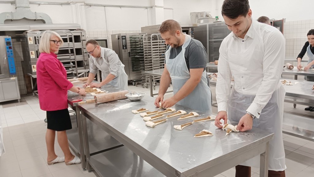 trzech mężczyzn w fartuchach za stołem piekarniczym przygotowuje Rogale Świętomarcińskie przed stołem kobieta obserwuje ich pracę