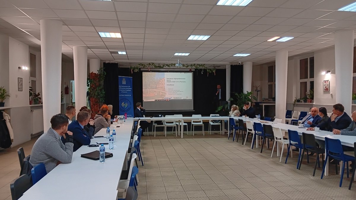 spotkanie w dużej sali, osoby siedzą przy stole, jedna osoba prezentuje na ekranie prezentację multimedialną