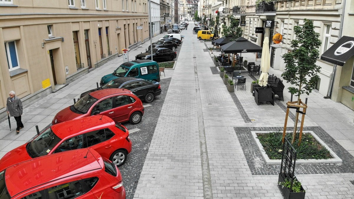 widom na wąską ulicę w zabudowie śródmiejskich kamienic, po lewej stronie zabarkowane ukośnie samochody, pod prawej ogródki gastronomiczne na chodniku