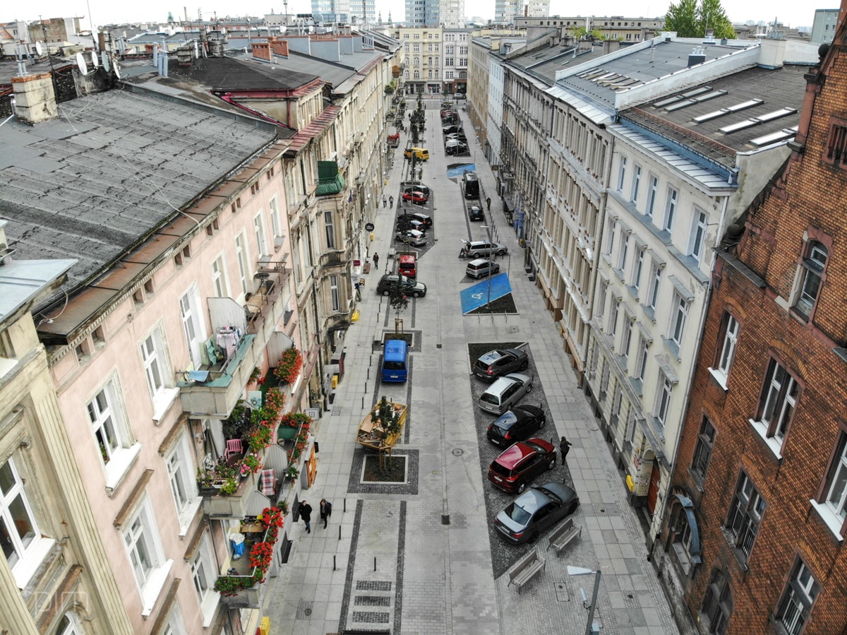 widok z dużej wysokości na wąską ulicę w zabudowie śródmiejskich kamienic, po prawej stronie samochody zaparkowane skośnie, po lewej kilka samochodów zaparkowanych równolegle do ulicy, pomiędzy ogródkami gastronomicznymi, są też miejsca parkingowe dla niepełnosprawnych - grafika artykułu