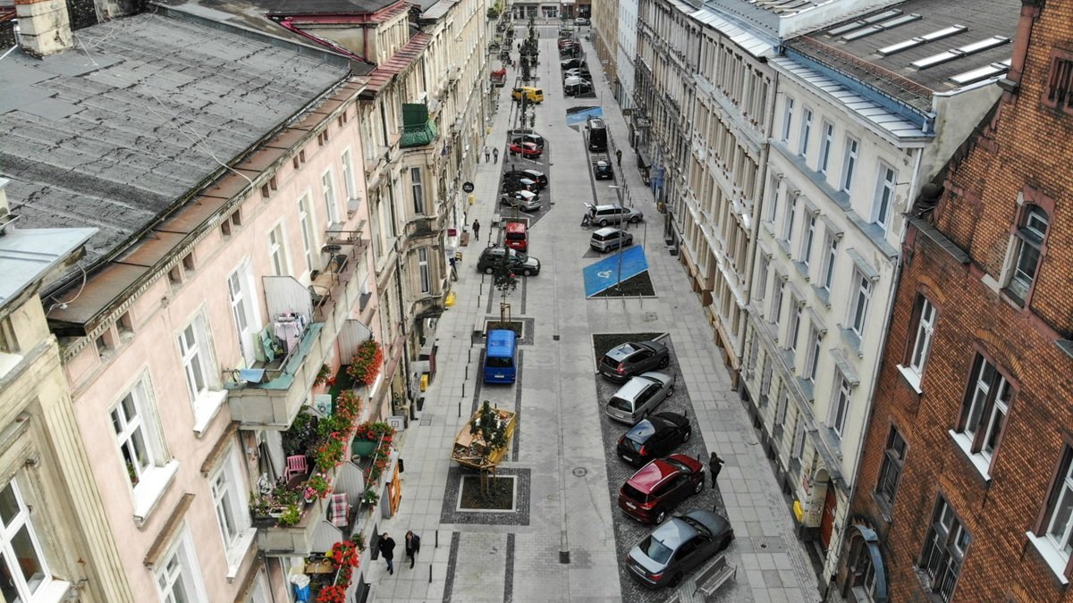 widok z dużej wysokości na wąską ulicę w zabudowie śródmiejskich kamienic, po prawej stronie samochody zaparkowane skośnie, po lewej kilka samochodów zaparkowanych równolegle do ulicy, pomiędzy ogródkami gastronomicznymi, są też miejsca parkingowe dla niepełnosprawnych