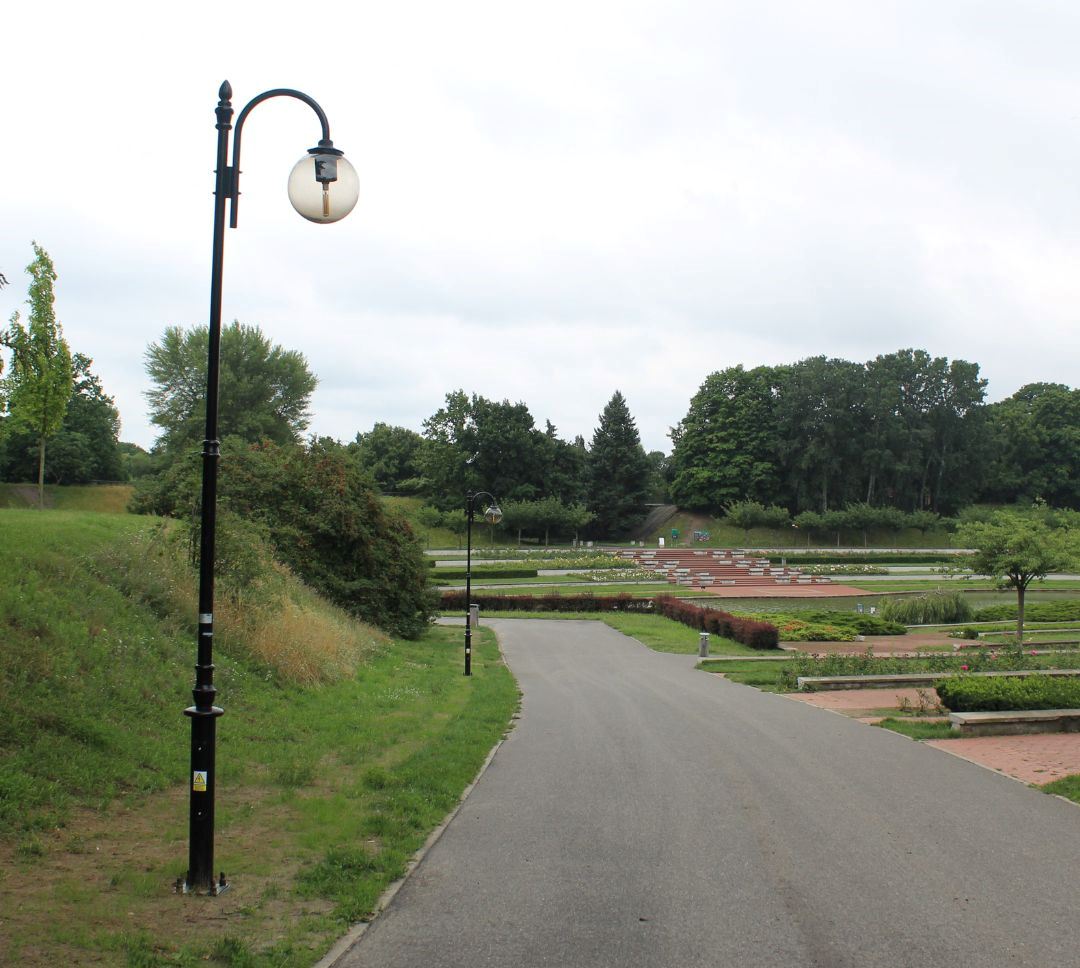 fragmnet parku z asfatową ścieżką wiodącą do oczka wodnego otoczonego ławkami, kwiatami, niską ziesilenią, przy alejce lampy uliczne, w dalszej części wysokie drzewa - grafika artykułu