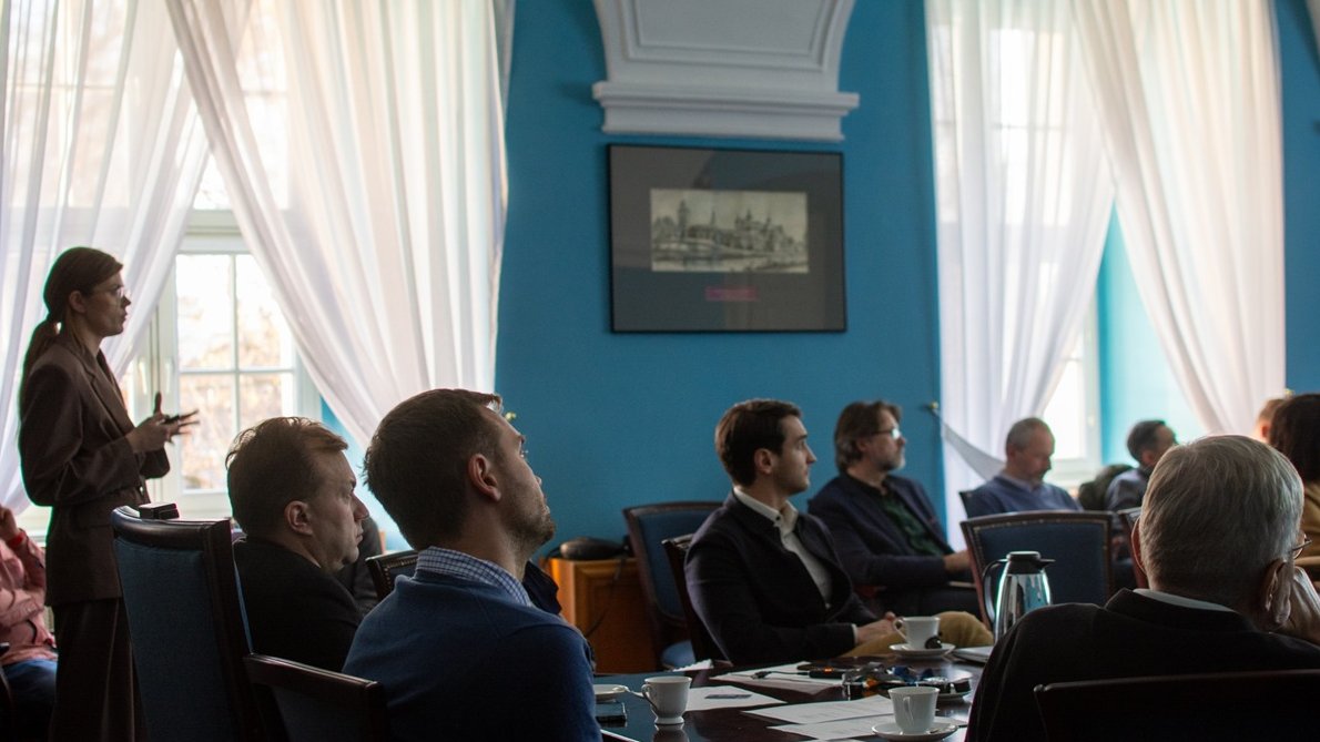 W sali konferencyjnej przy stole siedzi kilka osób, inne osoby siedzą na krzesłach za stołem, jedna osoba stoi i przedstawia prezentację