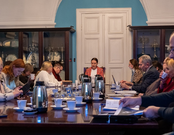 Przy stole konferencyjnym obraduje około kilkanaścioro osób, kobiet i mężczyzn w różnym wieku