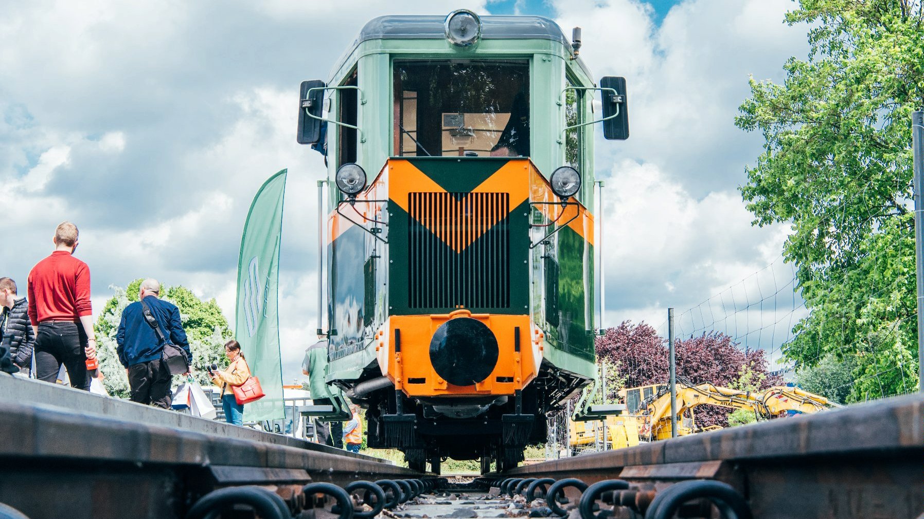 Widok na przód zielonej lokomotywy z żółtymi pasami. Obok pasażerowie kolejki stoją na stacji. - grafika artykułu
