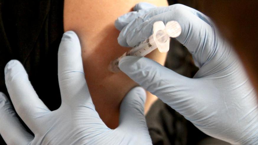 Na zdjęciu widać zbliżenie: dwie dłonie w medycznych rękawiczkach, które podają szczepionkę w ramię innej osoby - grafika artykułu