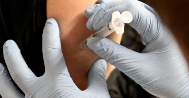 Na zdjęciu widać zbliżenie: dwie dłonie w medycznych rękawiczkach, które podają szczepionkę w ramię innej osoby