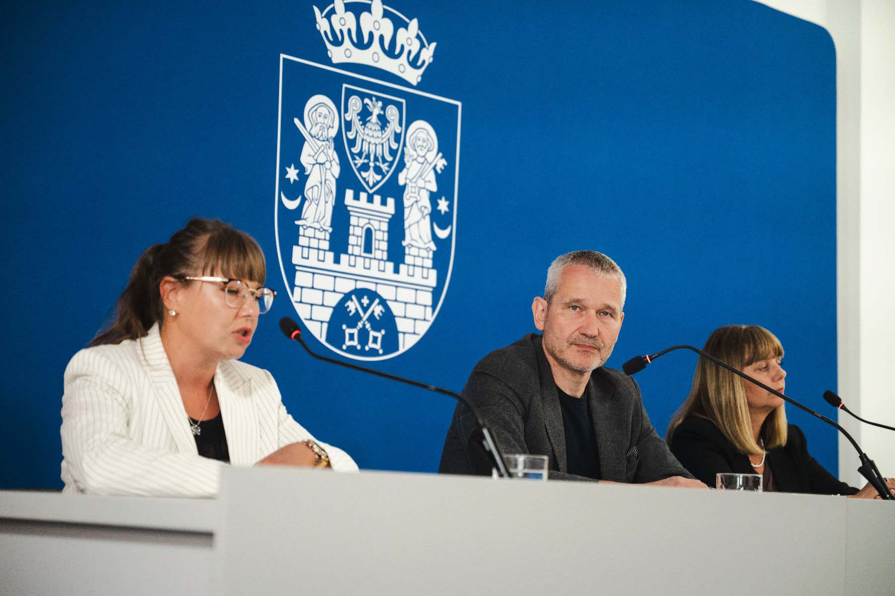 fot. Miasto Poznań trzy osoby na konferencji prasowej, w tle niebieska ściana i herb Poznania