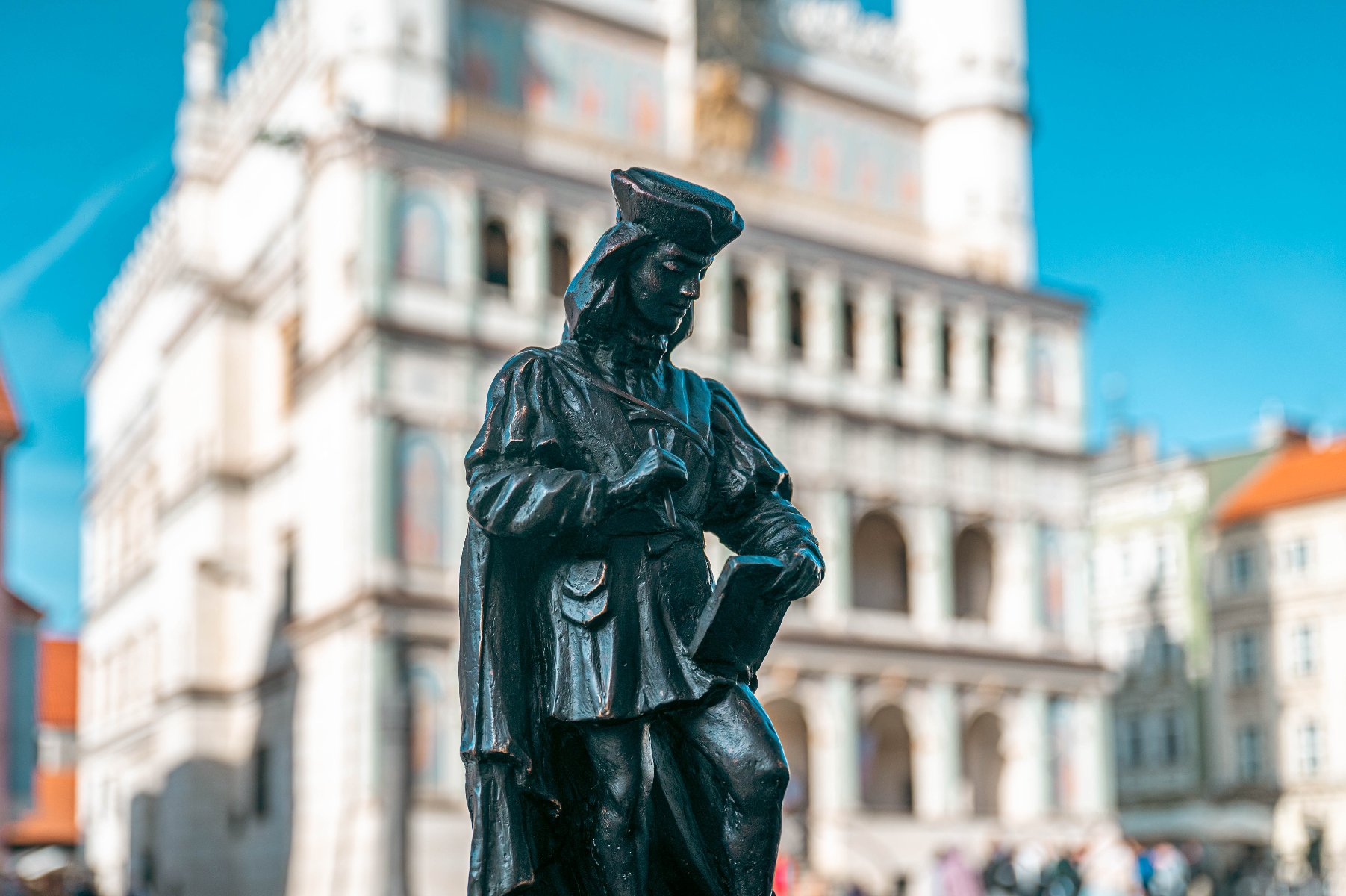 Znamy nominowanych do XXVII edycji Nagrody im. Jana Baptysty Quadro Czarna rzeźba w kształcie postaci w historycznym stroju, trzymającej zwój i pióro, na pierwszym planie. W tle, zamazany, biały renesansowy Ratusz w Poznaniu.