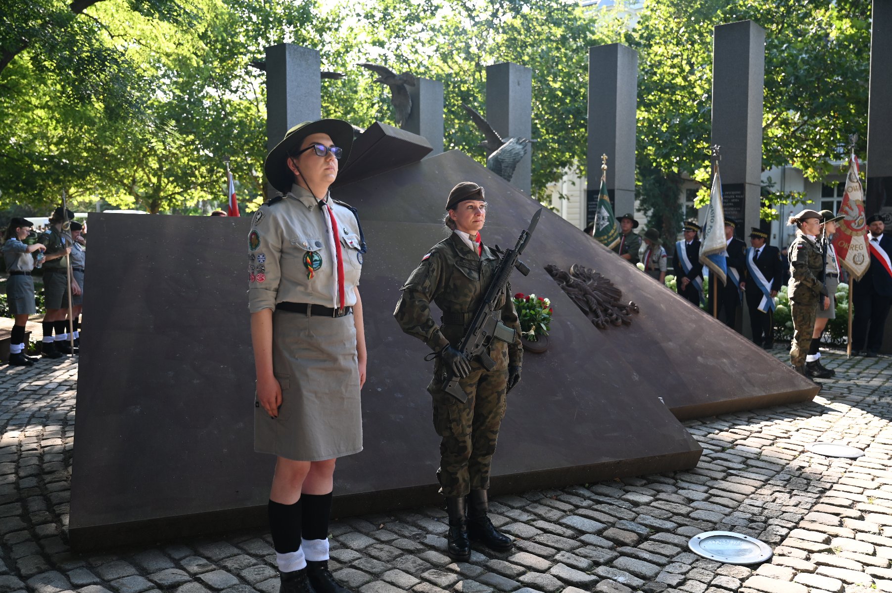 Równo 81 lat temu, 1 sierpnia 1944 roku wybuchła największa akcja zbrojna w okupowanej przez Niemców Europie - Powstanie Warszawskie Dwóch młodych ludzi w mundurach stoi na baczność przed pomnikiem. Po lewej harcerka w jasnym mundurze i kapeluszu, a po prawej żołnierka w mundurze polowym z karabinem i kwiatami. Za nimi widoczny jest masywny, ciemny pomnik, a w tle zieleń drzew i inne postacie w mundurach, oraz flagi. Scena wygląda jak uroczystość patriotyczna.