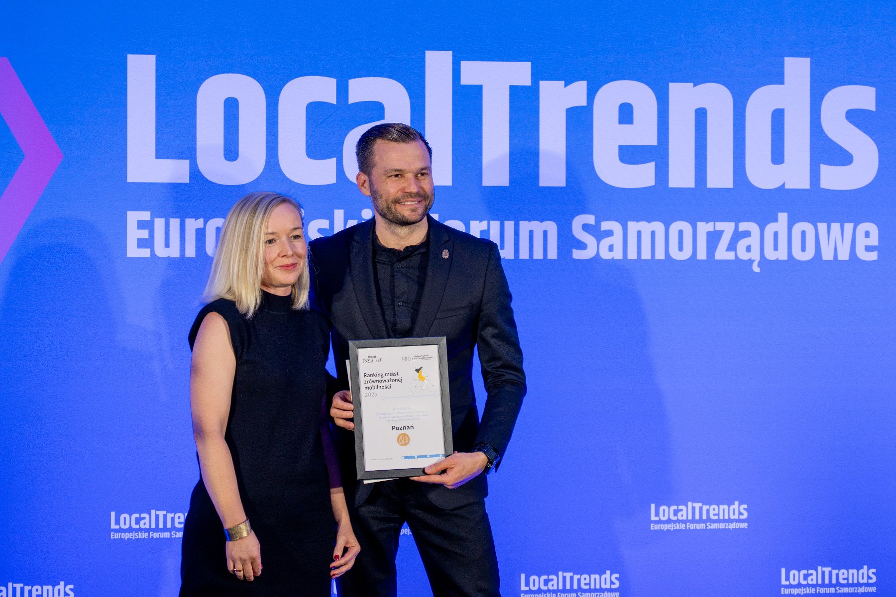 Poznań został doceniony za długofalową politykę w zakresie zrównoważonej mobilności oraz działania edukacyjne na rzecz czystego powietrza Kobieta i mężczyzna stoją na tle niebieskiego ekranu z logiem LocalTrends. Mężczyzna trzyma w rękach dyplom z napisem: "Poznań". Oboje uśmiechają się.