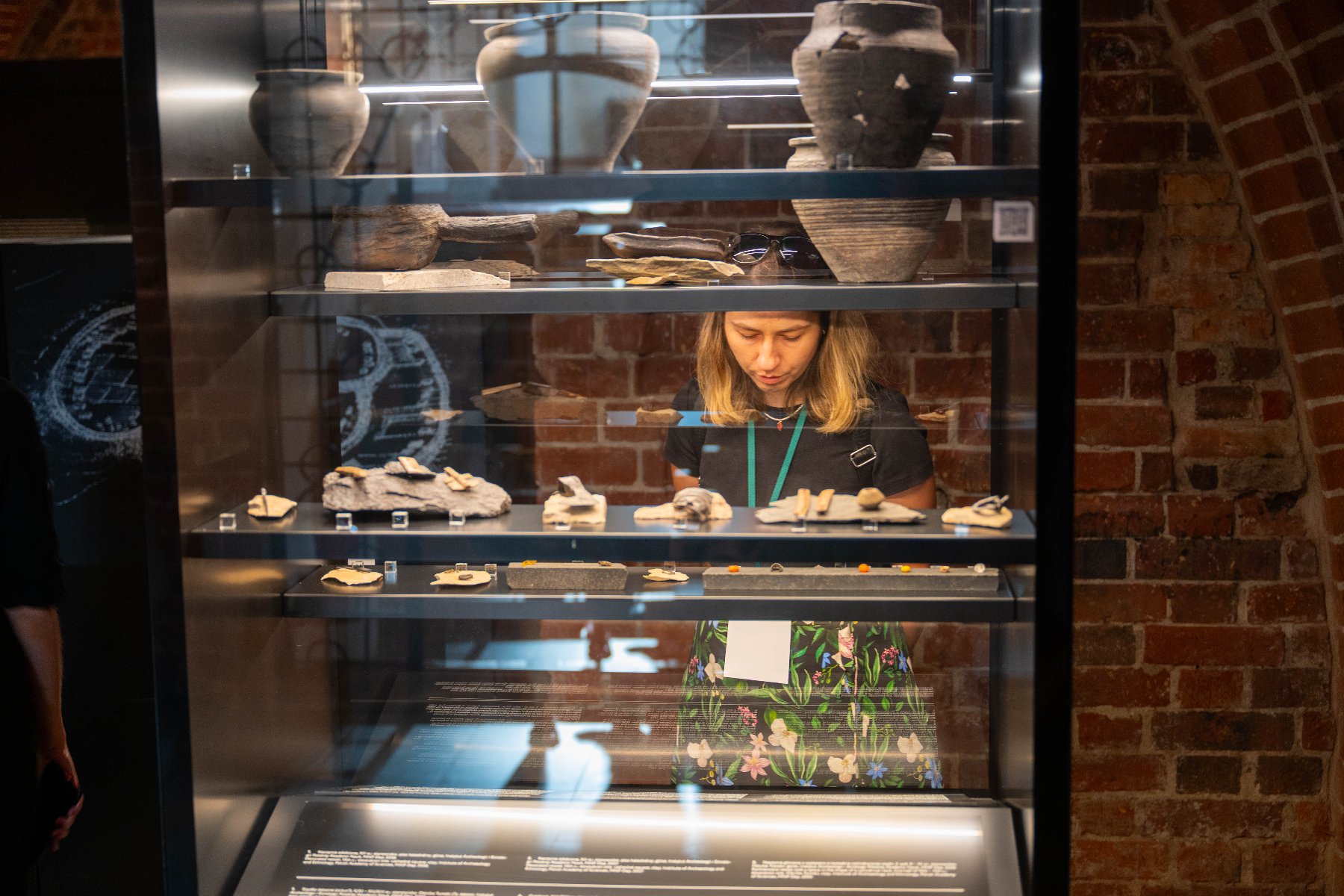 W Poznaniu trwa XI edycja Szkoły Letniej Polskiego Komitetu Narodowego Międzynarodowej Rady Ochrony Zabytków Kobieta ogląda szklaną gablotę, w której znajdują się ceramiczne naczynia i fragmenty prehistorycznych narzędzi. W tle ceglana ściana.