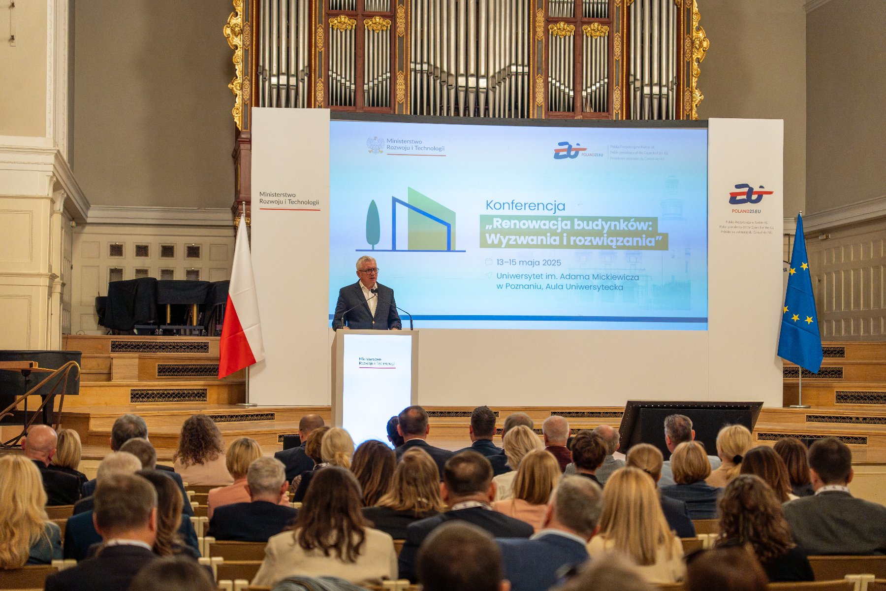 Wydarzenie poświęcone jest wyzwaniom i możliwościom związanym z poprawą efektywności energetycznej budynków oraz transformacją zasobów budowlanych w Polsce Konferencja w dużej auli z liczną publicznością. Prelegent stoi przy podium, przemawiając do zgromadzonych. Na dużym ekranie widnieje tytuł wydarzenia: "Renowacja budynków: Wyzwania i rozwiązania', organizowanego przez Ministerstwo Rozwoju i Technologii.