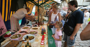 Zdjęcie przedstawia stoisko podczas jarmarku na Starym Rynku. Widać na nim kupujących ludzi i sprzedawcę oliwek