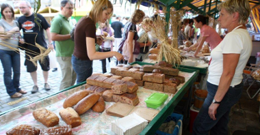 Zdjęcie przedstawia stoisko podczas jarmarku na Starym Rynku. Widać na nim kupujących ludzi i panie sprzedające chleb