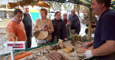 Zdjęcie przedstawia stoisko podczas jarmarku na Starym Rynku. Widać na nim kupujących ludzi i sprzedawcę ryb