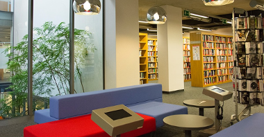 Zdjęcie przedstawia wnętrze biblioteki. Na pierwszym planie widać kanapy i stoliki, na dalszym - półki z książkami