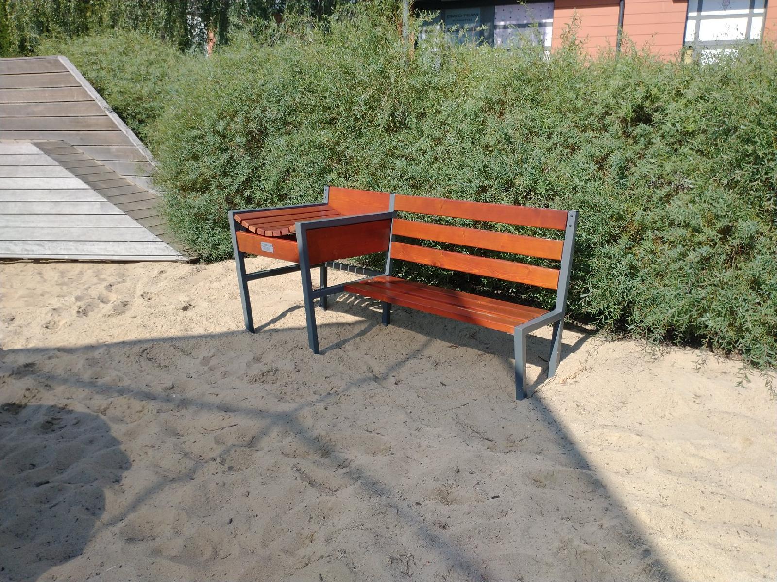 Na zdjęciu znajduje się ławka z zamontowanym przewijakiem dla dzieci. Ławka stoi na piasku, w tle widać zielone krzewy. - grafika artykułu