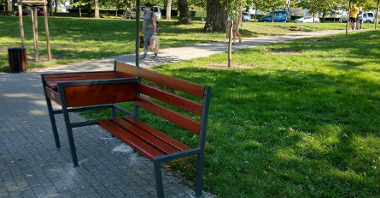 Na zdjęciu znajduje się ławka z zamontowanym przewijakiem dla dzieci. Ławka stoi w parkowej alei, na chodniku. W tle widać zieleń i spacerujących ludzi.