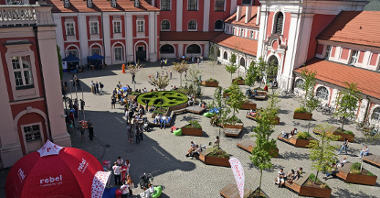 Zdjęcie przedstawia dziedziniec Urzędu Miasta Poznania. W miejscu tym wypoczywa wielu ludzi.