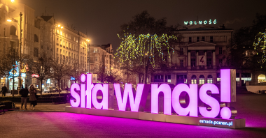 Zdjęcie przedstawia różowy, podświetlony neon z napisem "Siła w nas". Znajduje się on na placu Wolności. Zdjęcie zostało zrobione wieczorem.