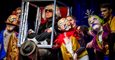Kadr ze spektaklu: w centrum aktor w pudełku-lektyce, wokół stłoczone kolorowe lalki