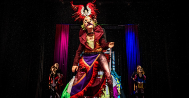 Kadr ze spektaklu: na scenie lalka Króla w kolorowych szatach i ogromnym czerwonym nakryciu głowy, w tle kulisy