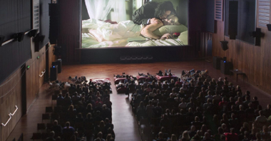 Zdjęcie przedstawia salę kinową. Na ekranie znajduje się kobieta leżąca w łóżku, trzymająca w dłoni różę oraz pochylający się nad nią mężczyzna. Na fotelach i pufach na przeciwko ekranu siedzą ludzie.