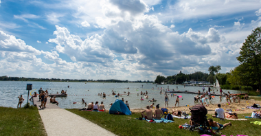 Zdjęcie przedstawia ludzi siedzących na plaży i kąpiących się w jeziorze.