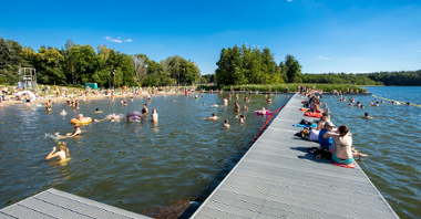 Zdjęcie przedstawia ludzi kąpiących się w jeziorze i siedzących na molo.