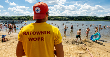 Zdjęcie przedstawia ludzi kąpiących się w jeziorze. Na pierwszym planie widać ratownika stojącego tyłem do aparatu i przodem do plaży.