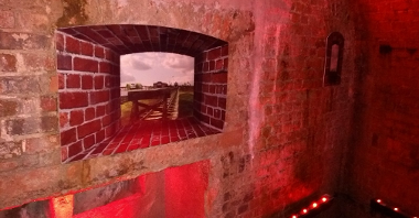 Zdjęcie przedstawia podziemia Kaponiery. Widać na nim ściany z cegły w czerwonym półmroku.