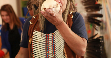Zdjęcie przedstawia mężczyznę w indiańskim pióropuszu trzymającego w ręku dużą muszlę.