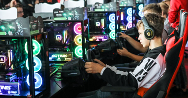 Zdjęcie przedstawia nastolatków grających w gry na symulatorze. W rękach trzymają urządzenie w formie kierownicy.