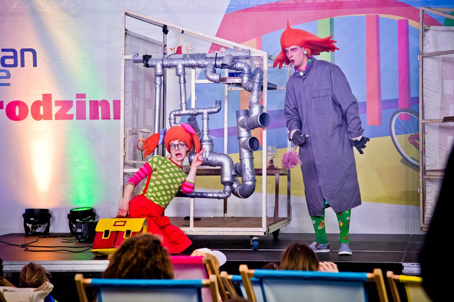 Na zdjęciu dwoje aktorów na scenie, sa kolorowo ubrani, mają zabawne czapki - grafika artykułu