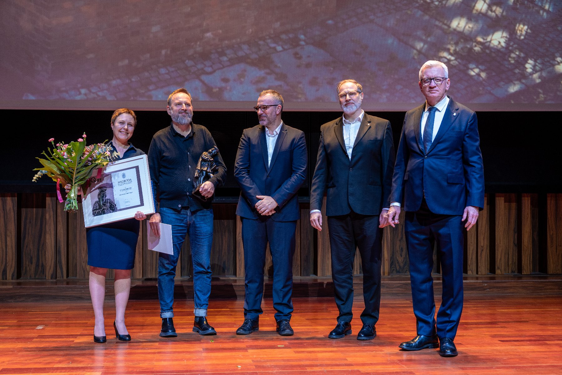 Food Fort Ostrów Tumski, zaprojektowany przez OZ Studio Projektowe, zwyciężył w XXVII edycji nagrody Jana Baptysty Quadro Pięć osób stoi na scenie podczas uroczystości wręczenia nagród. Kobieta po lewej, w niebieskiej sukience, trzyma bukiet kwiatów i oprawiony dyplom. Obok niej stoi mężczyzna w dżinsach i czarnym swetrze, trzymający statuetkę. Pozostali trzej mężczyźni są ubrani w garnitury.