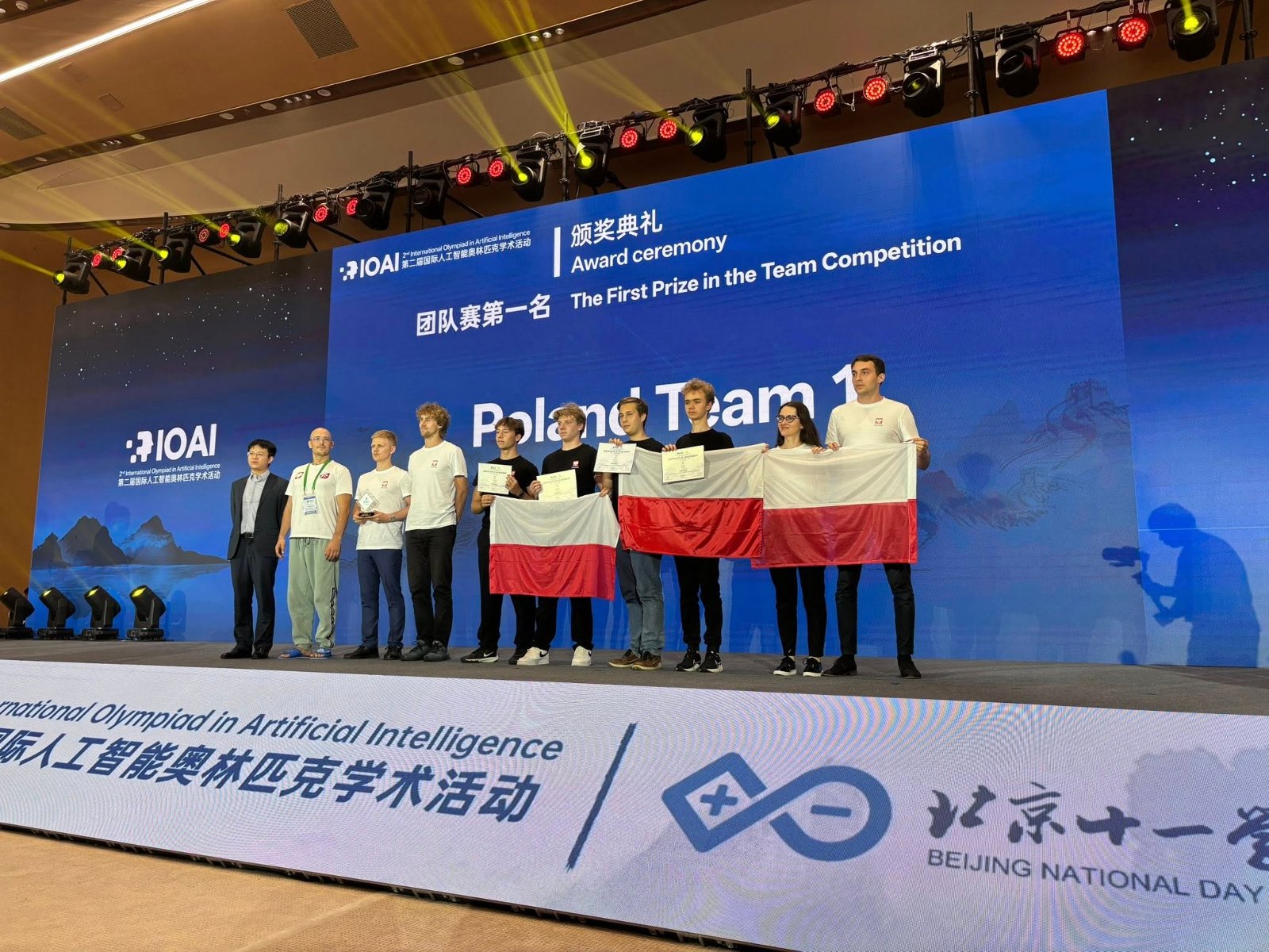 Reprezentacja Polski na Olimpiadzie Sztucznej Inteligencji w Pekinie. Fot. International Olympiad in Artificial Intelligence Polska reprezentacja pozuje na scenie z flagami. W tle napis: "pierwsza nagroda w kategorii drużynowej".