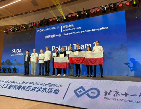 Reprezentacja Polski na Olimpiadzie Sztucznej Inteligencji w Pekinie. Fot. International Olympiad in Artificial Intelligence Polska reprezentacja pozuje na scenie z flagami. W tle napis: "pierwsza nagroda w kategorii drużynowej".