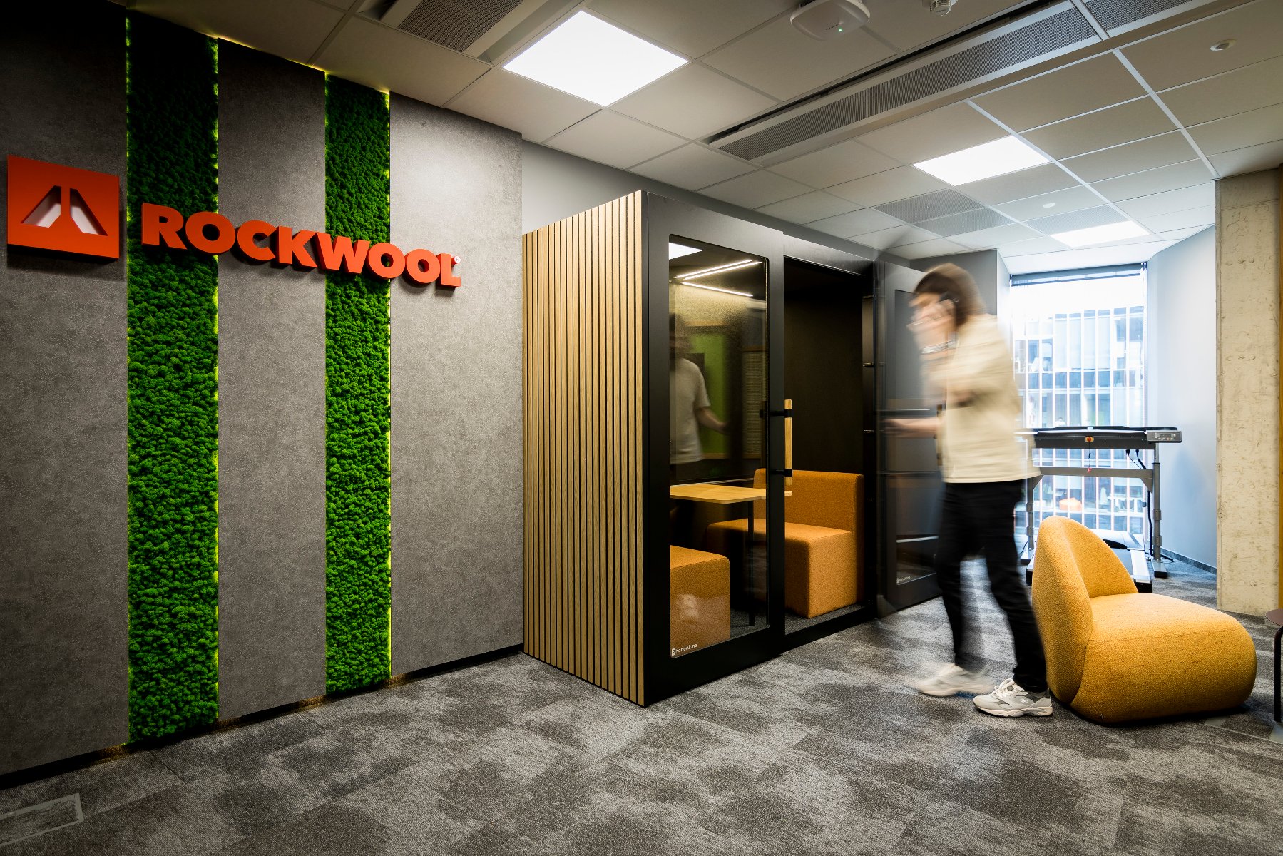 Poznański oddział ROCKWOOL świętuje 9. rocznicę Strefa ciszy na korytarzu biurowym. Na ścianie z pasem zieleni wisi logo ROCKWOOL.