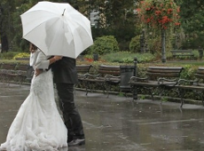 A newly married couple is standing together in a park on a rainy day. The bride is wearing a long, elegant white wedding dress with a flowing train, while the groom is dressed in a dark suit. They are holding each other closely under a large white umbrella, which protects them from the falling rain. Photo: CC BY-SA