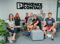 Phoenix Contact Wielkopolska is one of the largest employers on the local markets in Nowy Tomyśl, Poznań and Tarnobrzeg. A smiling group of people sit on couches in the office. A Phoenix Contact logo hangs on the wall.