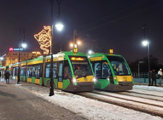 At a snowy tram stop at night, two modern green-and-yellow trams are standing side by side on the tracks. Streetlights illuminate the area, and one of the lampposts is decorated with a glowing Christmas angel light. Snow covers the ground and the rails. A few pedestrians in winter clothing are visible nearby. The scene feels calm, wintry, and festive.