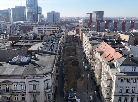 The photo shows a city street undergoing extensive construction work, likely for a tramway or road renovation. In the background, modern office towers and high-rises are visible, while historic multi-story tenement buildings line both sides of the street.