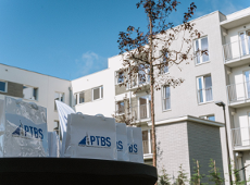 Kilka białych toreb z logo "PTBS' ustawionych na czarnym stole. W tle nowoczesny budynek mieszkalny z wieloma piętrami, balkonami i oknami. Widać również drzewo o rzadkich liściach oraz jasne niebo z kilkoma chmurami. Sceneria sugeruje wydarzenie związane z działalnością deweloperską lub prezentacją projektu mieszkaniowego.