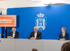 Na zdjęciu dwóch mężczyzn i kobieta za stołem konferencyjnym, w tle niebieska ścianka z herbem miasta