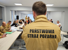 Na zdjęciu widać ludzi podczas szkolenia w sali. Na pierwszym planie jest osoba w żółtej koszulce z napisem Państwowa Straż Pożarna. Osoba ta stoi tyłem do obiektywu. W tle zdjęcia widać inne osoby, które siedzą przy stołach.