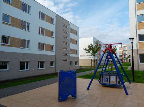 Zakończyła się budowa II etapu inwestycji mieszkaniowej realizowanej w ramach miejskiego programu "POZnań - i zamieszkaj" Plac zabaw z czerwoną i niebieską huśtawką przystosowaną dla osób z niepełnosprawnościami na tle nowoczesnych, jasnych budynków mieszkalnych. Na jednym z budynków widoczny napis: "Elżbiety Zawackiej". Po lewej stronie niebieski panel edukacyjny.
