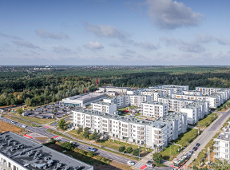Zdjęcie jest zrobione z lotu ptaka. Z przodu widać kompleks budynków w białym kolorze. Obok kompleksu widać ulice i parking. W tle zdjęcia widać duży obszar zieleni.