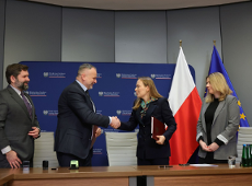 Zdjęcie przedstawia czwórkę ludzi - dwóch mężczyzn i dwie kobiety - stojących przy stole. Mężczyzna i kobieta znajdujący się pośrodku podają sobie ręce. W tle widać flagi Polski i Unii Europejskiej oraz ściankę z nazwą ministerstwa.