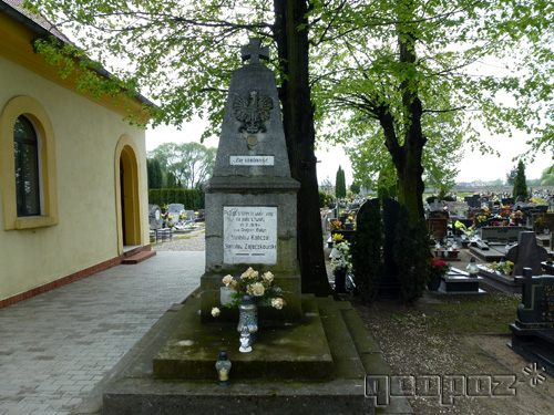 Pomnik z orłem - godłem. Na grobie ustawiony bukiet z róż. Po lewej stronie kaplica na cmentarzu. Po prawej wysokie drzewa i inne groby.
